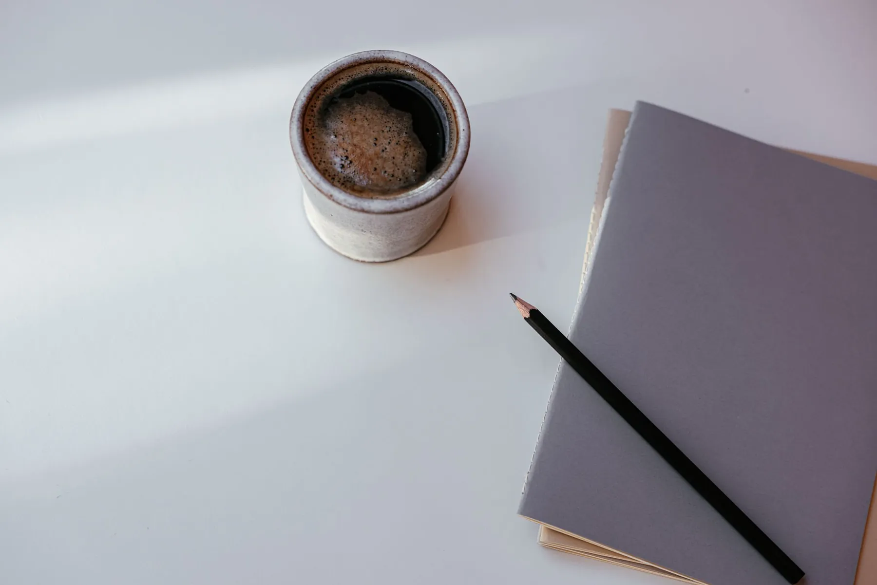 A cup of coffee beside a notebook and a pencil on a white desk.