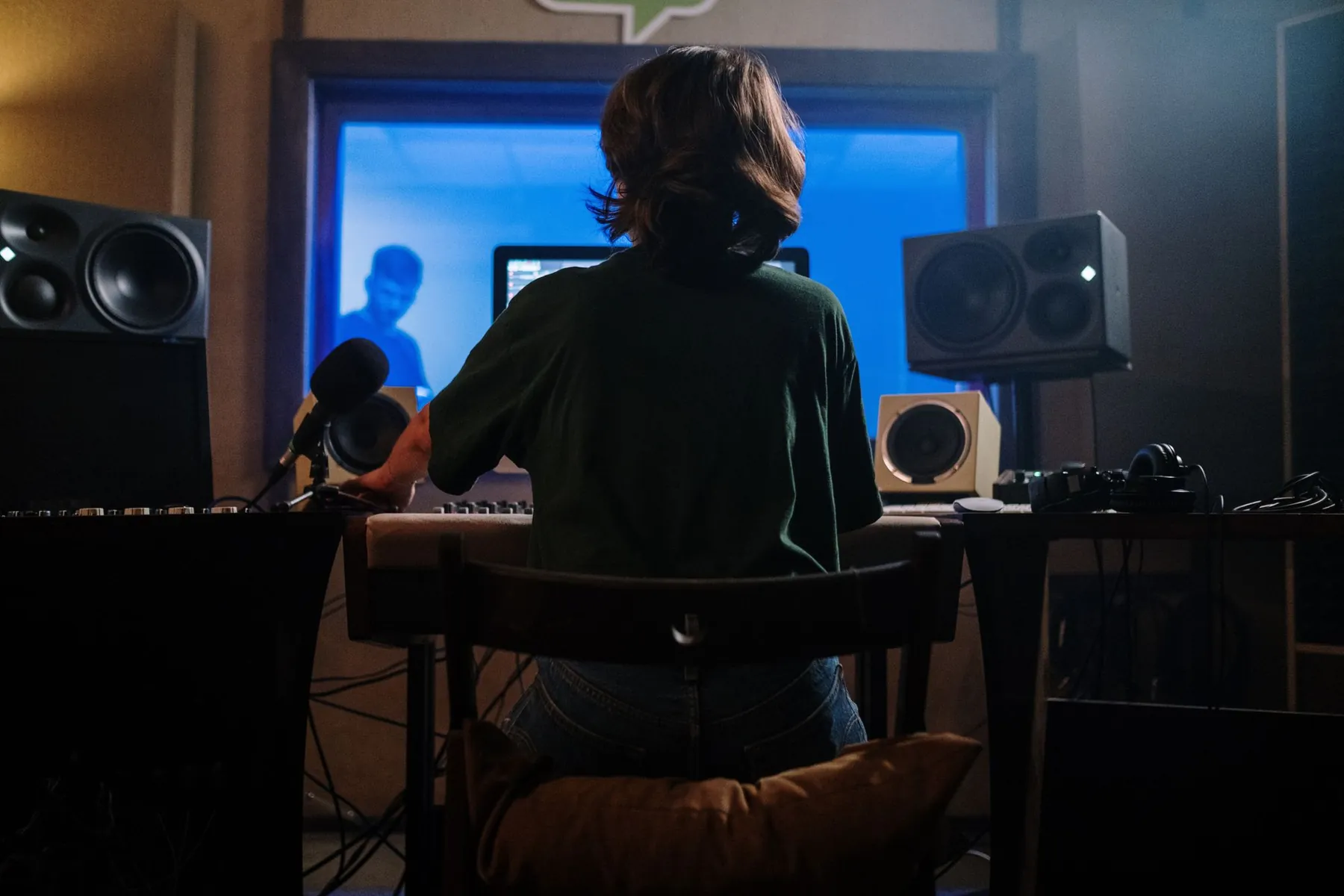A woman at the studio desk, facing the vocal booth during a session.